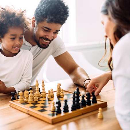 dark skinned, male presenting person with a dark skinned child and woman playing chess