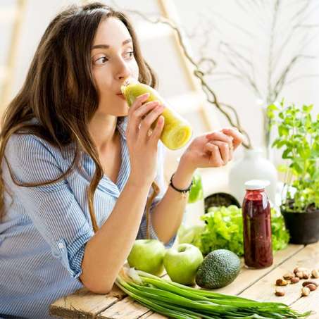 light-skinned, female presenting person drinking green juice leaning against counter filled with green vegetables and fruit