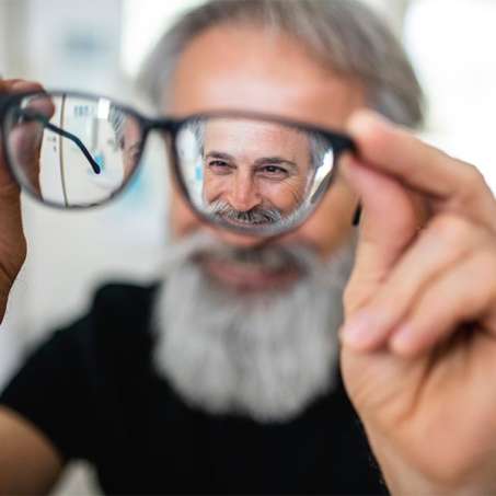 light-skinned, older grey haired and beard male presenting person holding glasses away from face and looking through them