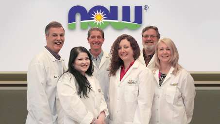 six NOW workers of various race and genders wearing lab coats in front of NOW logo smiling