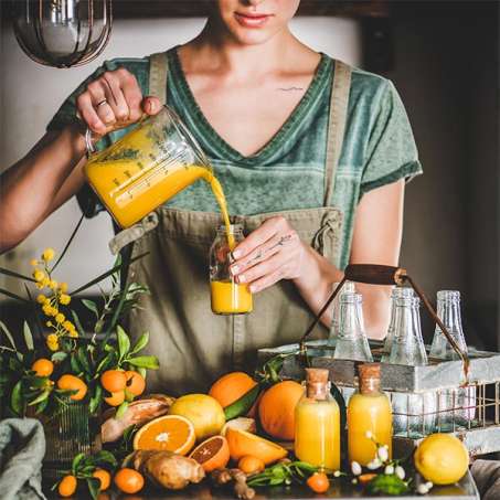 light-skinned, female presenting person pouring orange juice into glass bottle surrounded by orange fruit on counter