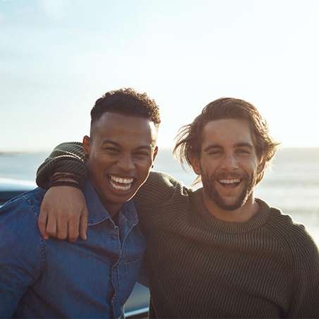 one dark skinned male presenting person and one light skinned male presenting person smiling embracing and smiling at the camera 