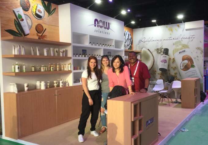 4 adults of various races and genders smiling in front of a NOW trade show display