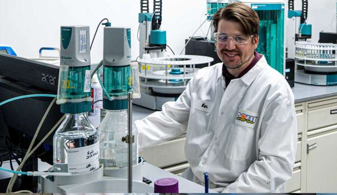 light skin male presenting person wearing a white lab coat sitting next to various testing and lab equipment