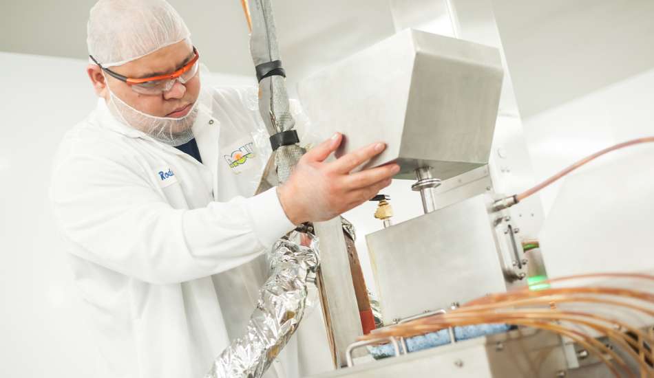 light skinned male presenting person working on a manufacturing machine wearing a hairnet, beard-net, and white lab coat 