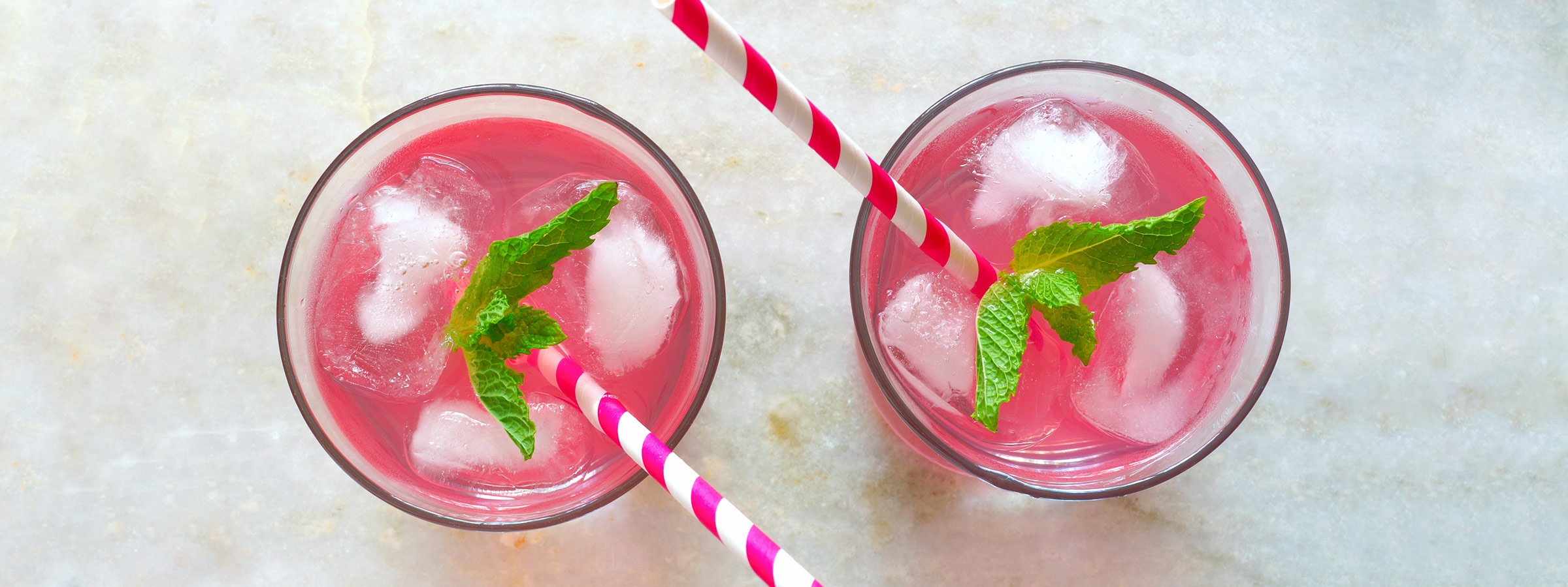 two pink drinks in an ice cold glass with ice cubes, straws and mint leaf
