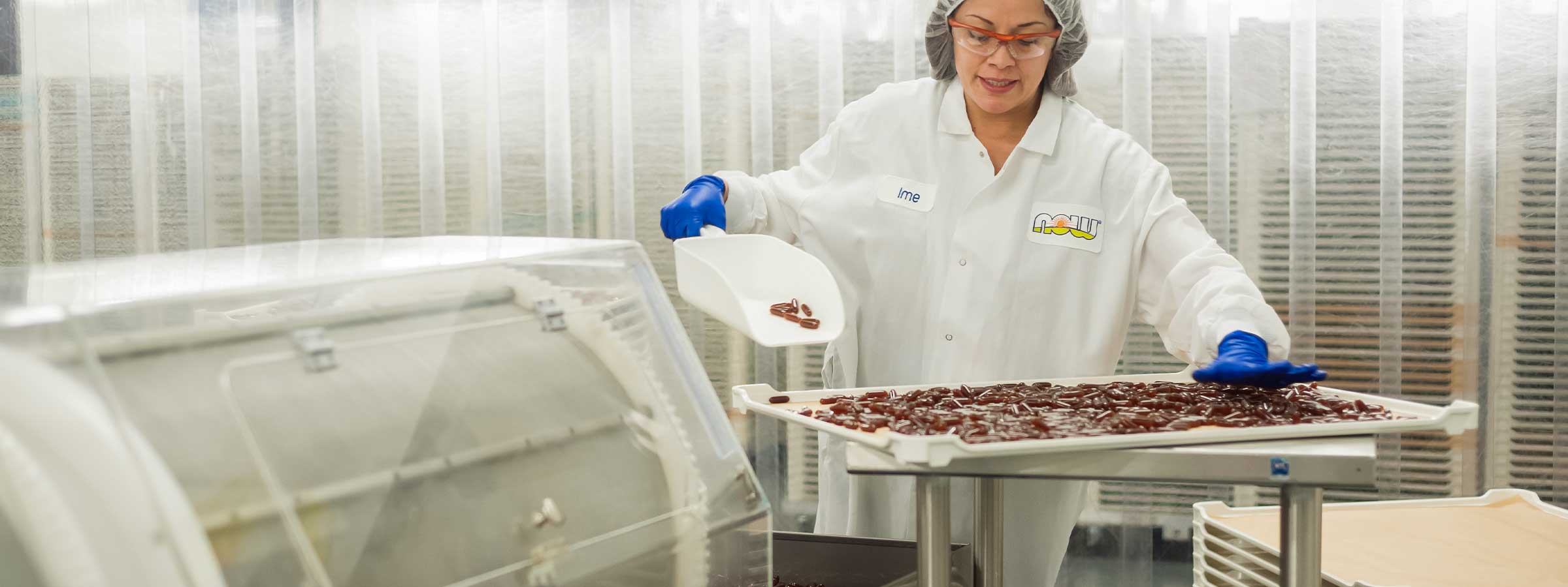 medium skin female presenting person wearing lab coat and blue gloves shoveling red softgels onto a metal tray