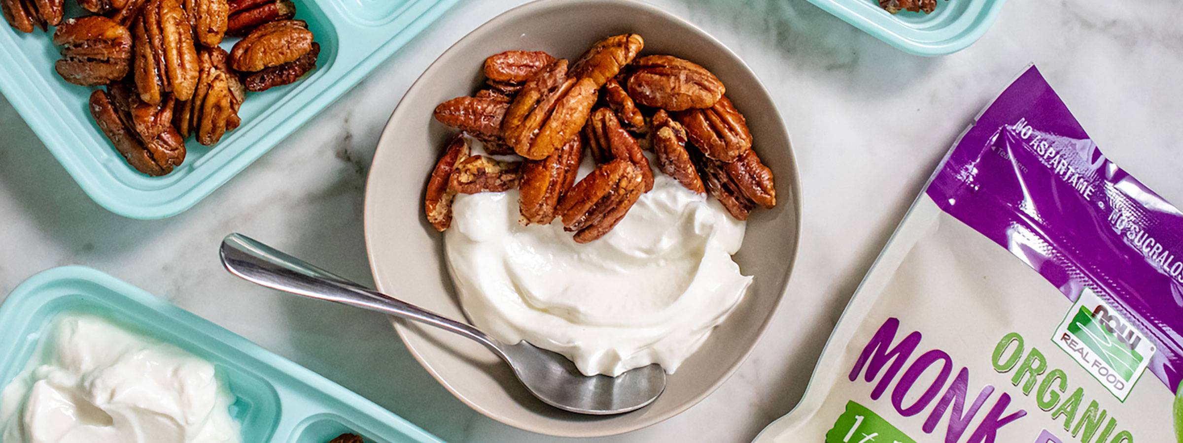 overhead image of white bowl with candied pecans on one side and creamy yogurt on the other, partial view of aqua colored trays surrounding the bowl with same contents and a bag of NOW Monk Fruit with Erythritol