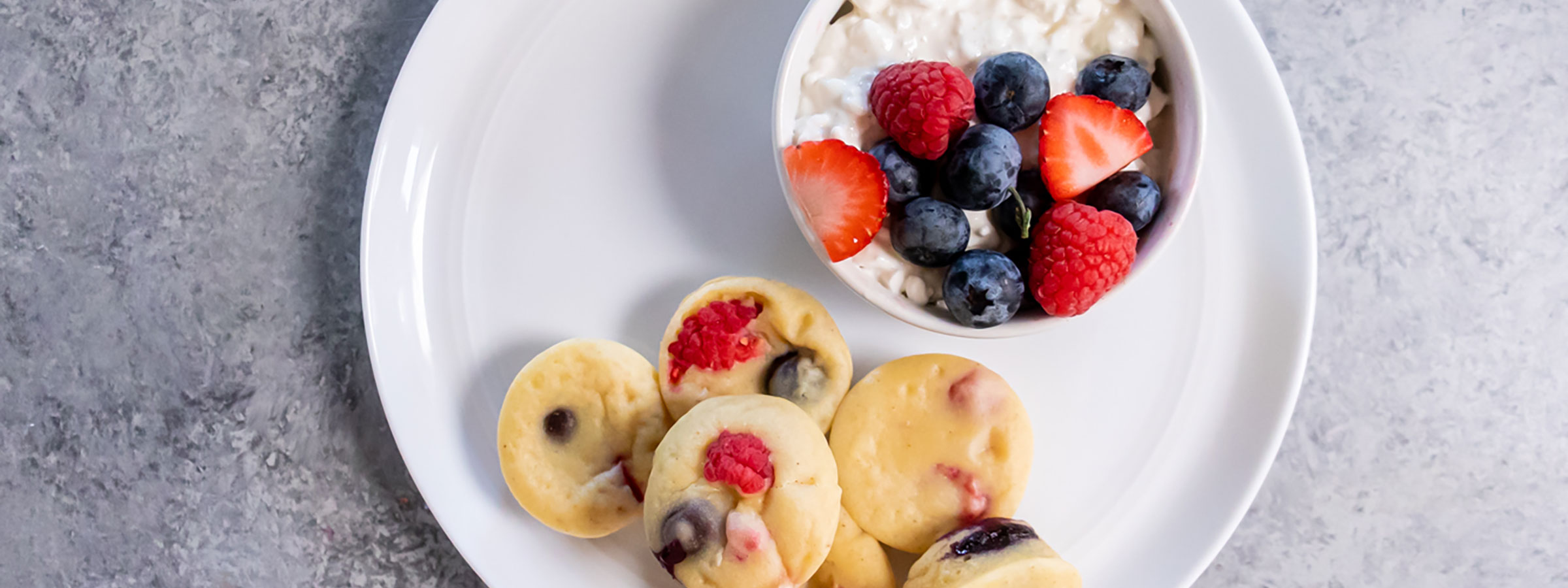 overhead view of five small protein pancake bites on a white plate with a small cup of mixed berries and yogurt