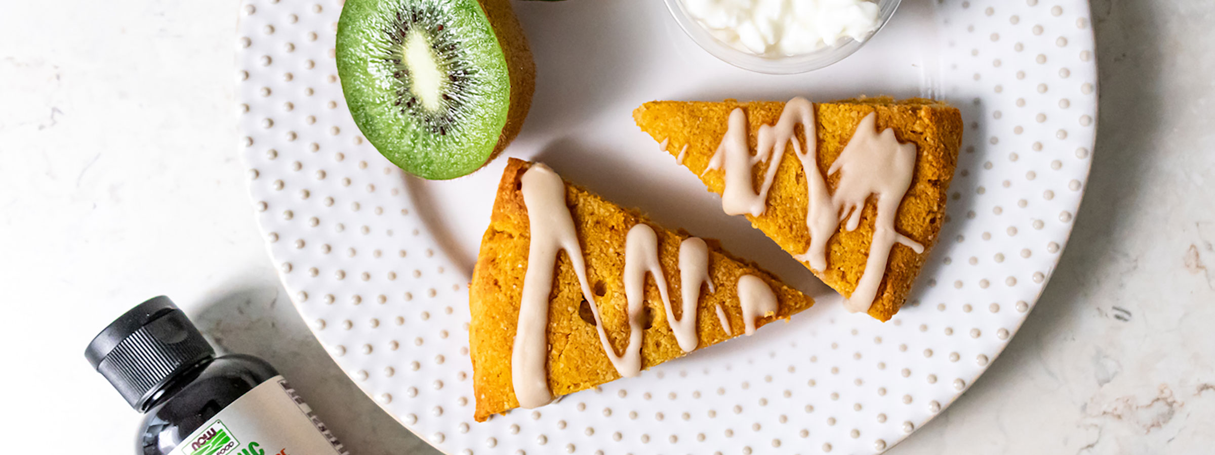 two scones with pumpkin glaze on a white plate which also has kiwi slices and yogurt on it viewed from the top