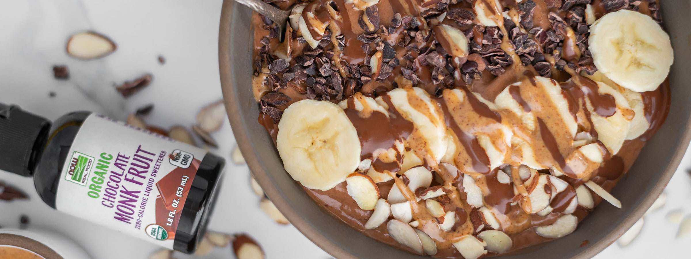 brown bowl filled with double chocolate smoothie bowl mixture including sliced bananas on top and a bottle of NOW Chocolate Monk Fruit sweetener laying to the left
