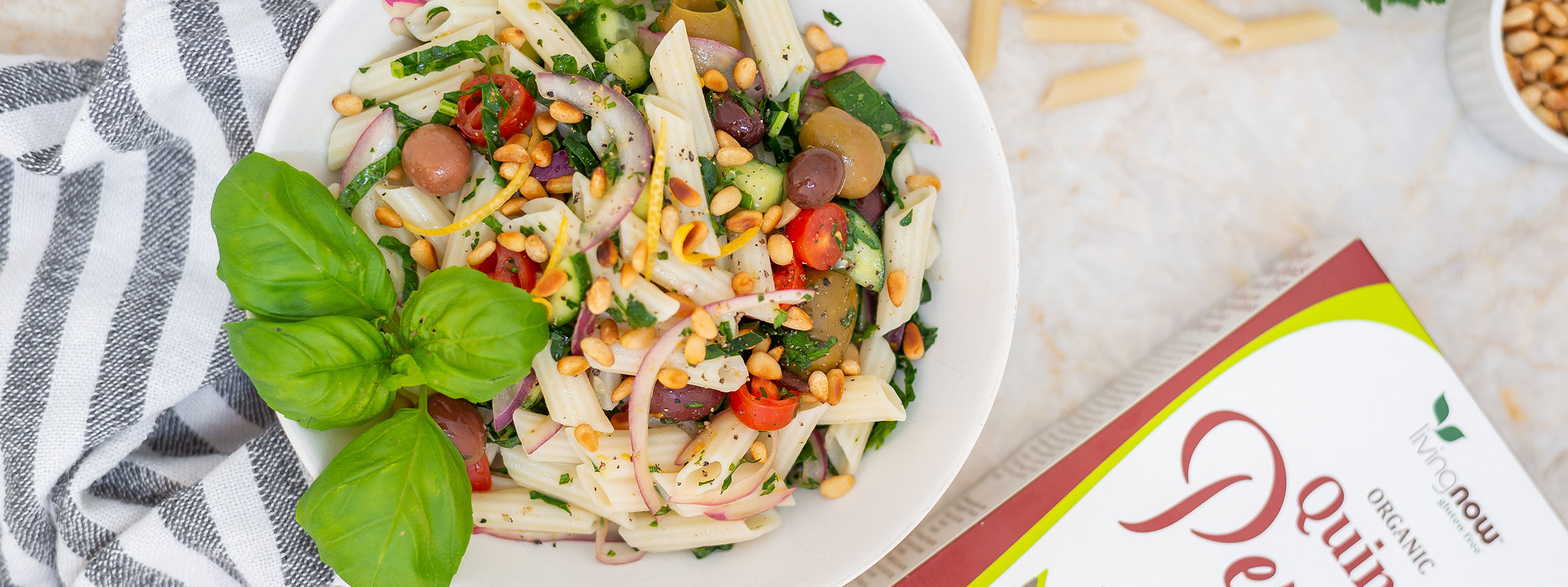 colorful bowl of penne pasta salad from an overhead view with a blue and white napkin on the left side and box of NOW Penne Pasta on the right