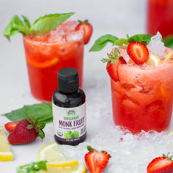 two glasses of strawberry basil lemondade with sliced strawberry on top sitting on a table of crushed ice with a bottle of NOW Monk Fruit sweetener next to the glasses