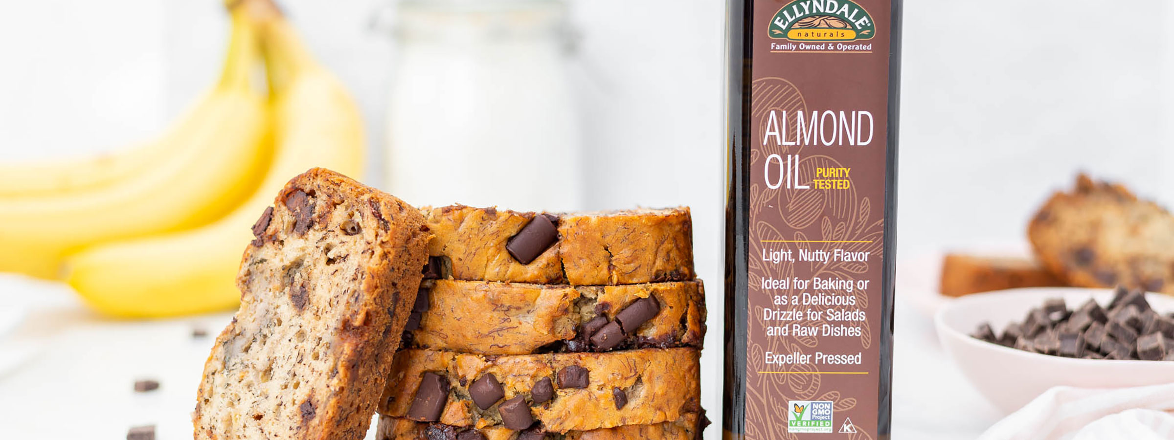 chocolate banana bread slices stacked on a table with one slice leaning against the stack, a bottle of NOW Ellyndale Almond Oil to the right of the stack and bananas and chocolate chips in the background