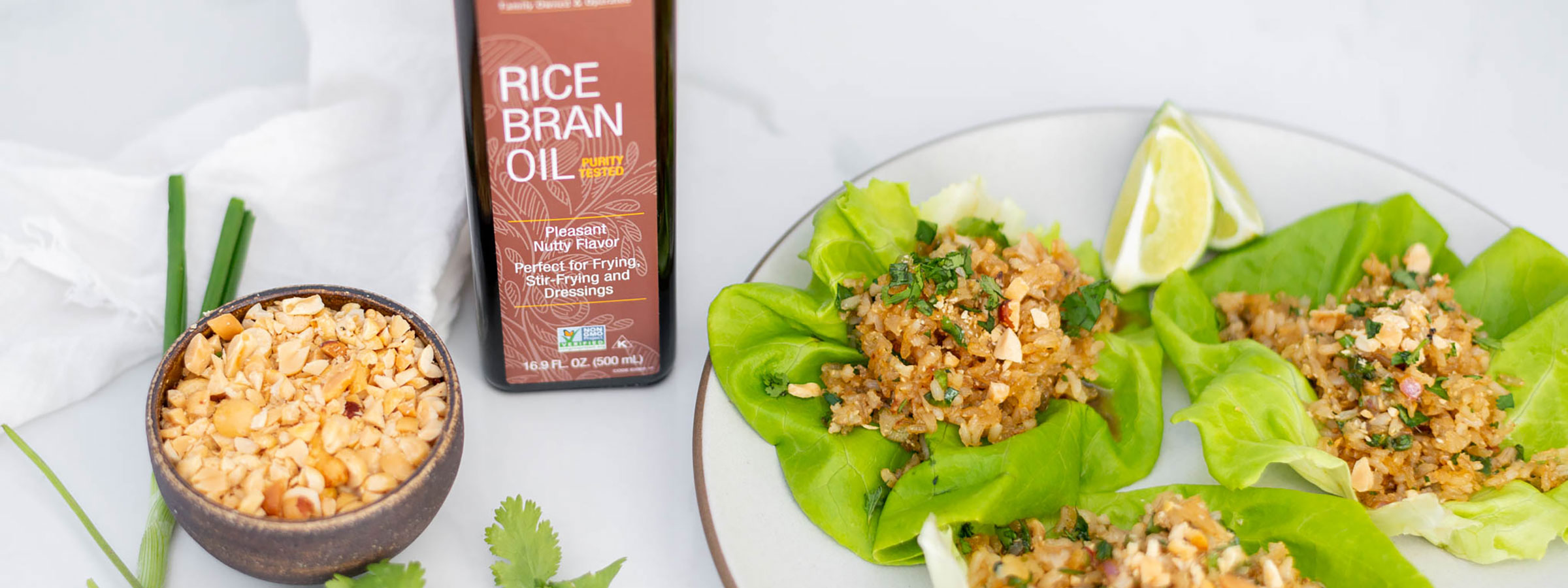 a white plate with three open lettuce wraps and two lemon wedges, a bottle of NOW Ellyndale Rice Bran Oil to the left of the plate and a small wooden bowl of nuts and dried fruit to the far left