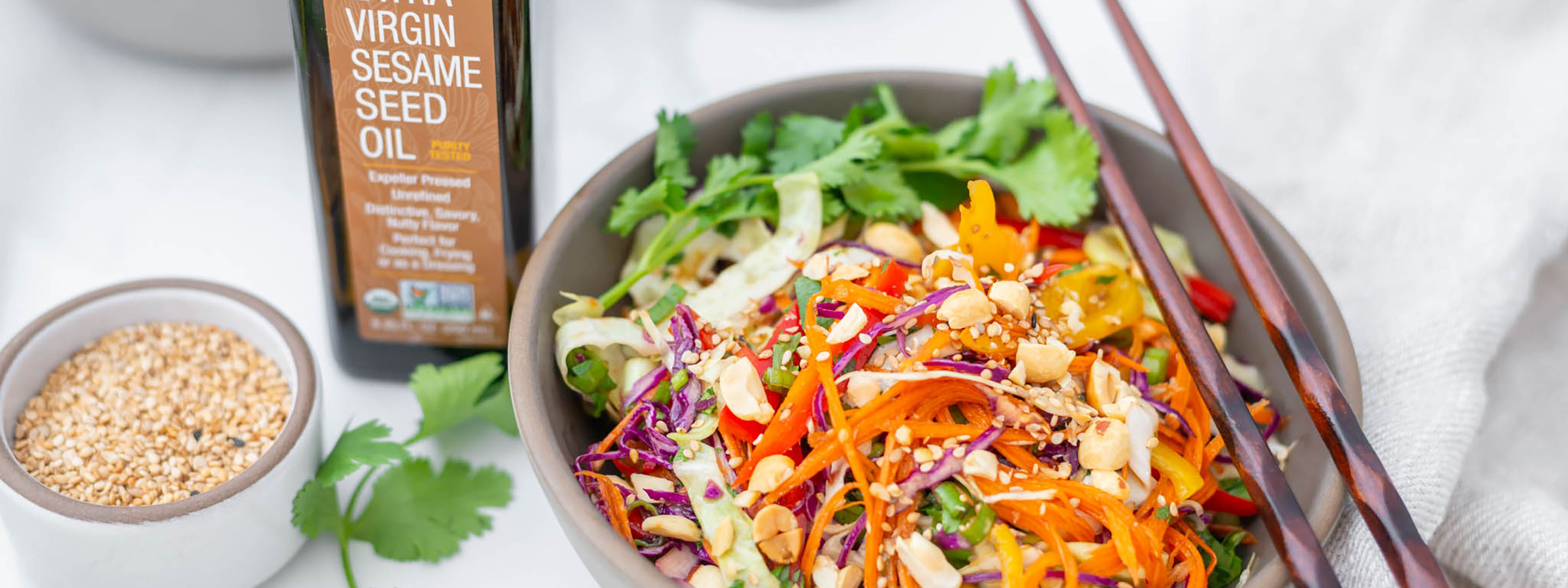 colorful bowl of Thai salad with chopsticks resting on the bowl, a bottle of NOW Sesame Seed Oil to the left and a small bowl of sesame seeds to the left