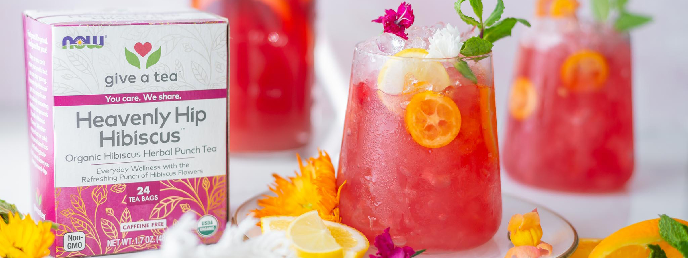 glass filled with a hibiscus tropical fruit punch dink surrounded by NOW Heavenly Hibiscus Tea and various fruits and flower garnishes