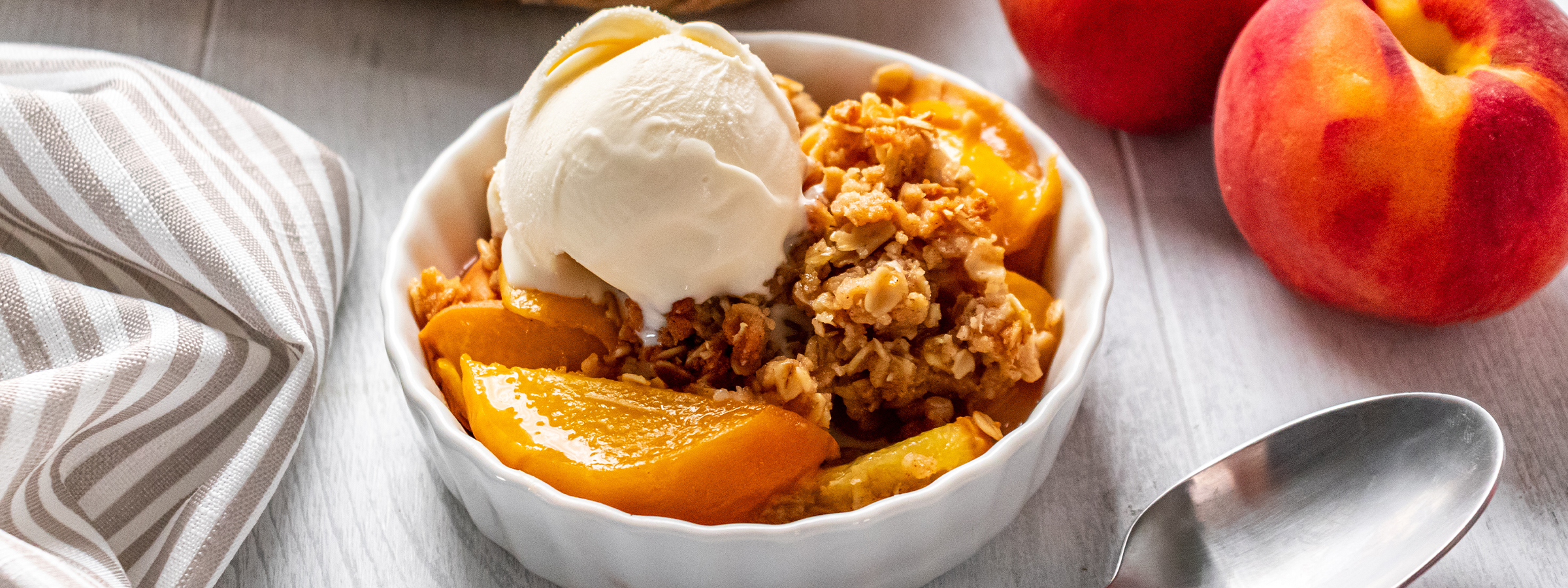 top view of bowl with peach crumble with an oat topping and a dollop of ice cream next to two peaches