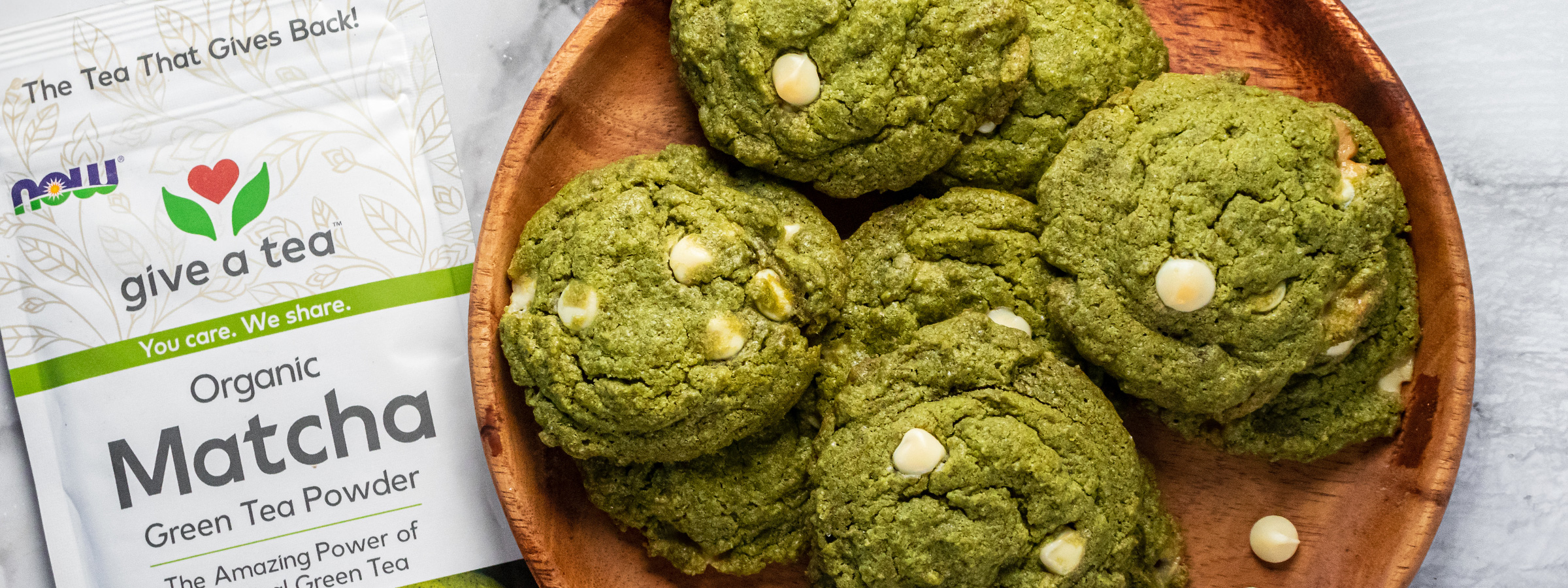 top view of matcha white chocolate cookies on wooden plate next to bag of NOW Matcha tea