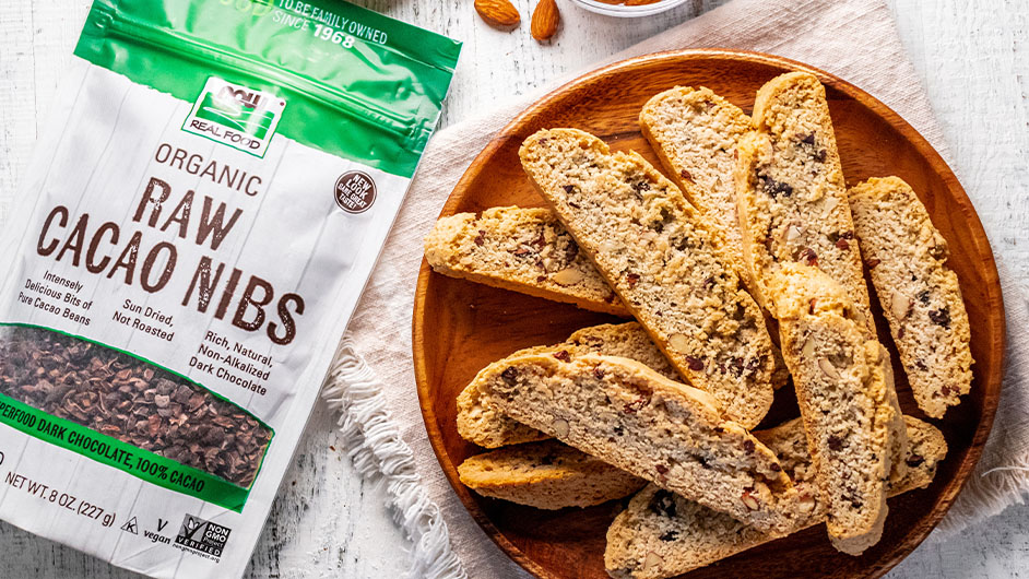 top view of Almond and Cacao Nib Biscotti on brown plate next to bag of NOW Raw Cacao Nibs