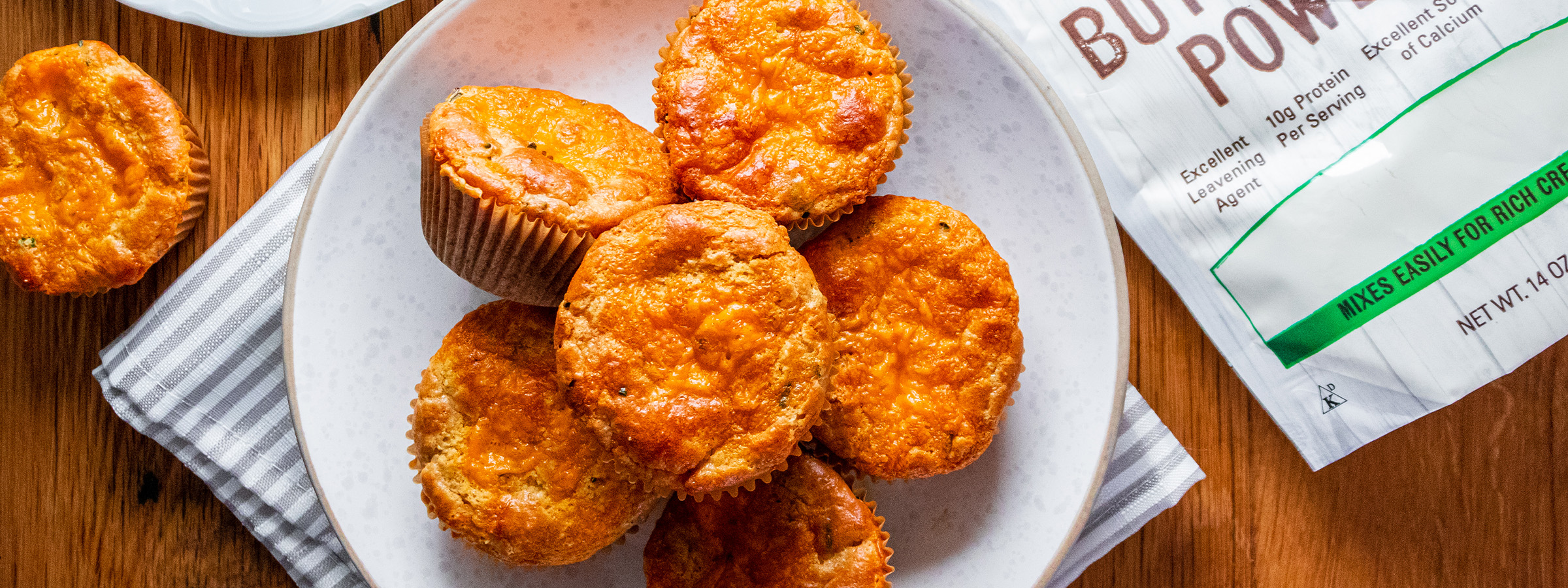 top view of Buttermilk, Cheddar and Herb Muffins on a white plate next to a bag of NOW Buttermilk Powder