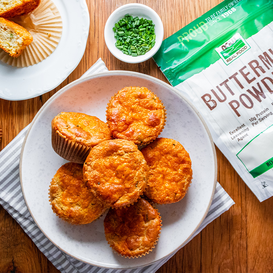 top view of Buttermilk, Cheddar and Herb Muffins on a white plate next to a bag of NOW Buttermilk Powder