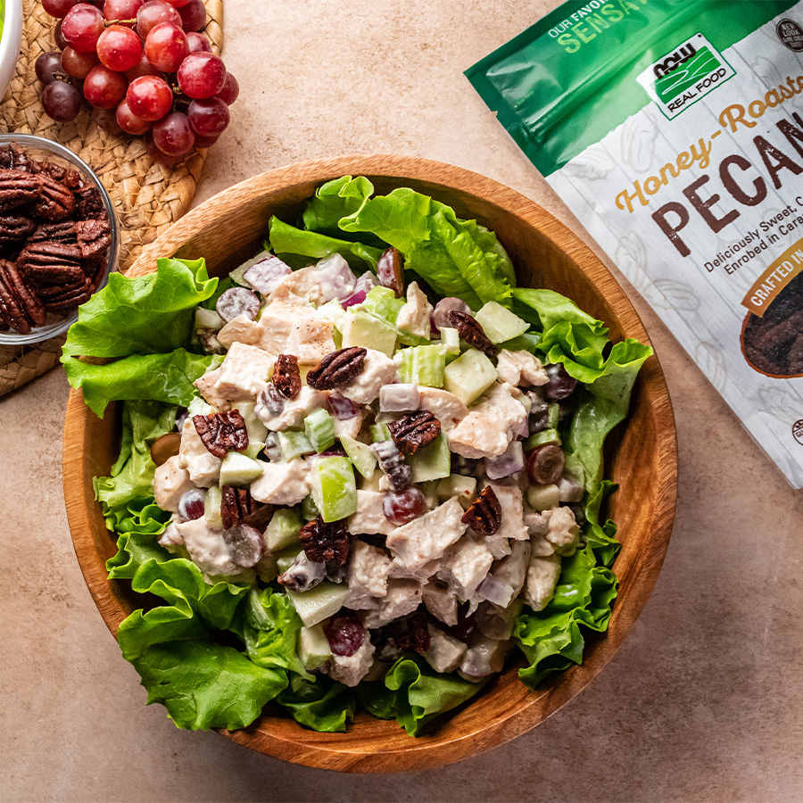 top view of Waldor Chicken Salad in a wooden bowl next to some grapes and NOW Honey Roasted Pecans