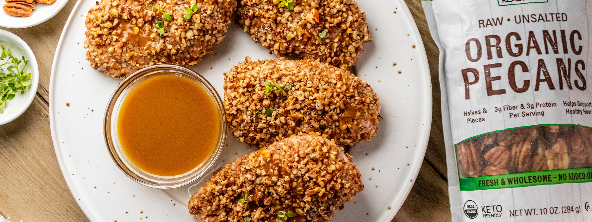 top view of Pecan Herb Crusted Chicken with maple dijon sauce on a white plate next to a bag of NOW Organic Pecans