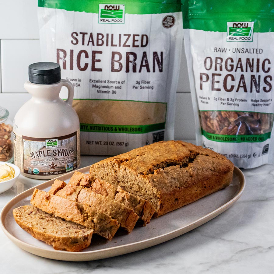 white plate with slices of Rice Bran Banana Nut Bread with bottle of Maple syrup, bag of NOW Real Food Stabilized Rice Bran and bag NOW Real Food Organic Pecans