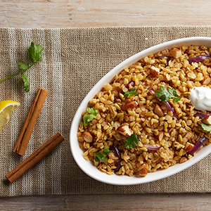 A white ceramic bowl on a brown placemat holds a serving of Lebanese Freekeh with Lentils & Onions.