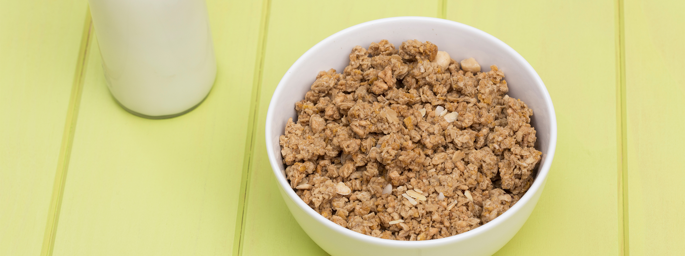 A white ceramic bowl on a yellow wooden table holding NOW Millet Granola.