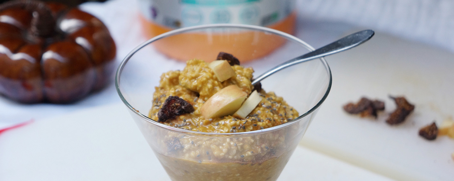 Round glass bowl of cooked steel cut oats and a spoon with the handle of it sticking out on the right, topped with chia seeds, dried fig bits and apple chunks. On the white counter in the blurred background from left to right is a brown ceramic pumpkin, the bottom portion of a NOW Sports bottle likely of whey protein and chopped figs.