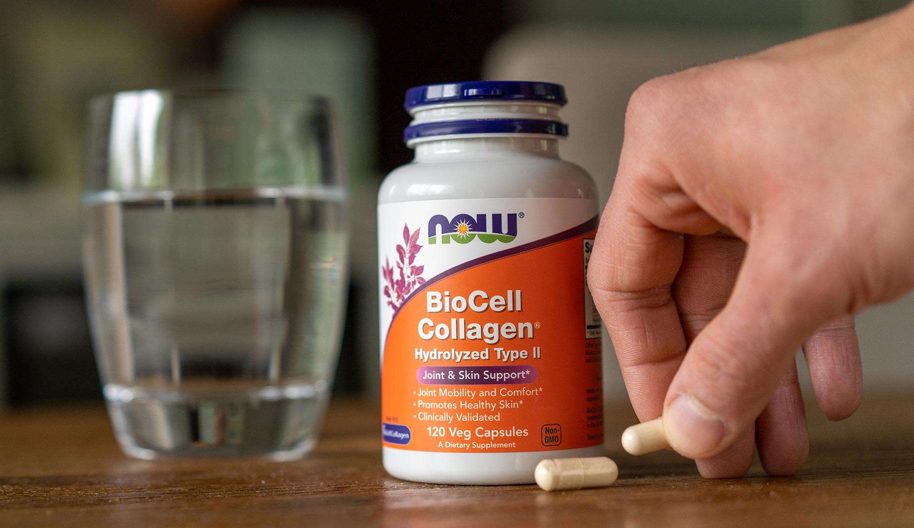 glass of water next to a bottle or NOW BioCell Collagen with hand picking up a pill