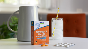 Omega-3 Fish Oil Gummy product orange box with white ceramic pitcher and glass of ice water in background on white counter with two orange chewables in foreground.