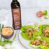 a white plate with three open lettuce wraps and two lemon wedges, a bottle of NOW Ellyndale Rice Bran Oil to the left of the plate and a small wooden bowl of nuts and dried fruit to the far left