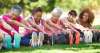 Group of multi-age and multi-race female presenting people on the grass, legs outstretched, leaning forward touching toes