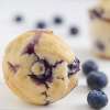 A moist Blueberry Muffin lays on a table facing the camera. Several blueberries are also placed.