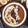 white bowl of Chocolate smoothy with slices of bananas, chia seeds, coconut and chocolate shavings