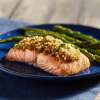 A dark blue plate on a wooden table holds a serving of Manuka Honey & Sesame Ginger-Crusted Salmon