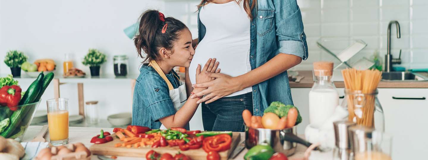 young girl kissing her pregnant mothers belly while preparing food in the kitchen