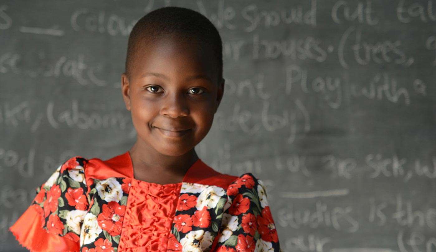 A dark-skinned fem-presenting child stands in front of a blackboard wearing a red dress patterned with white flowers.