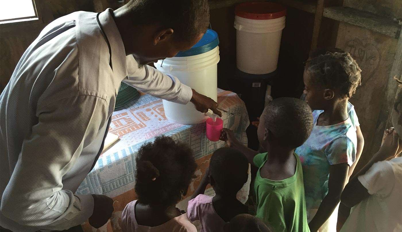 Four dark-skinned children stand around a water dispenser with a dark-skinned male-presenting person pouring water into a pink cup.