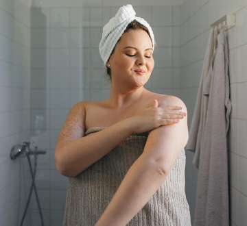 Light skinned female presenting person in a towel applying body lotion after a shower