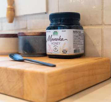jar of NOW Real Food Manuka Honey on a wooden block with a spoon sitting in front of it