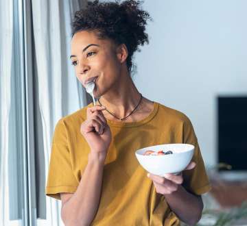 dark skinned female presenting person holding a bowl of food looking out a window with a spoon in her mouth