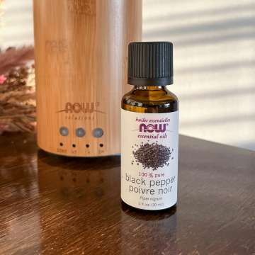 NOW Black Pepper Essential Oil bottle in front of a NOW Bamboo diffuser on a brown table with dried flowers in the background