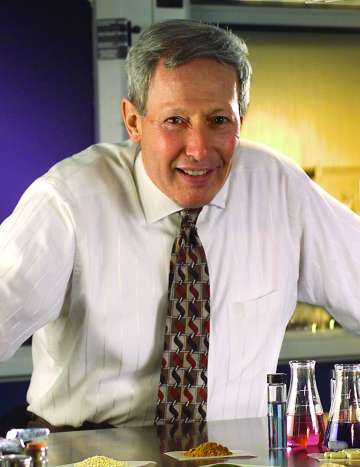 A light-skinned male-presenting person with silver hair, white dress shirt, and patterned tie smiles while leaning on a metal table.