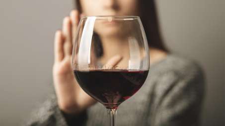 glass of red wine in foreground with light skinned female presenting person in the background putting up her hand in refusal of the wine