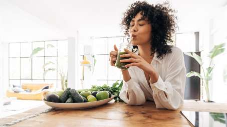 Dark-skinned female presenting person drinking a green smoothie through a straw. Sitting at a wooden table with a bowl full of green fruits and Vegetables on the table