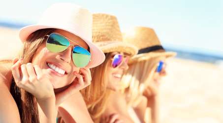 three female presenting persons in sunglasses and hats at the beach smiling at the camera
